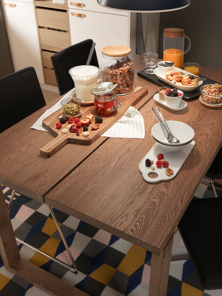 A wooden dining table set for breakfast with a large serving board of fresh fruits, jars of granola and honey, small bowls, and a pitcher of orange juice, over a colorful geometric rug.