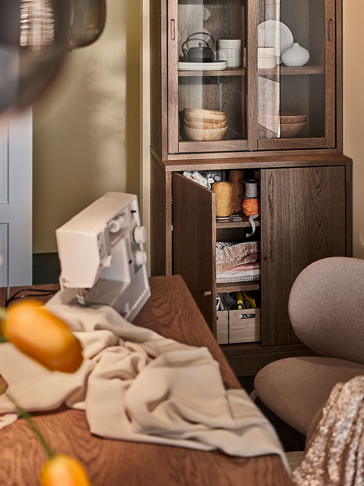 A sewing machine sits on a wooden table with beige fabric in the foreground, next to a tall wooden cabinet with glass doors on top and open lower doors showing craft supplies.