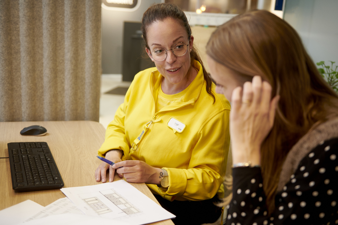 An IKEA employee in a yellow uniform and glasses assisting a customer with a design floor plan.